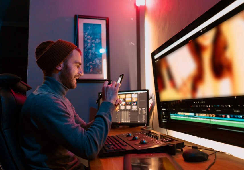 Attractive Male Video Editor Works with Footage or Video on His Personal Computer and having a break communicating on his smartphone. He Works in Creative Office Studio or home. Neon lights